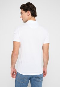 Biała koszulka polo z krótkim rękawem o gładkiej fakturze i klasycznym kołnierzykiem, noszona z niebieskimi dżinsami, ukazująca widok z tyłu.