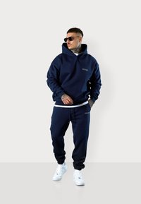 Marinefarbener Hoodie mit Fronttasche und Logo, kombiniert mit passender Jogginghose. Abgerundet mit weißen Sneakern. Weicher Stoff, lockere Passform und Kapuze.