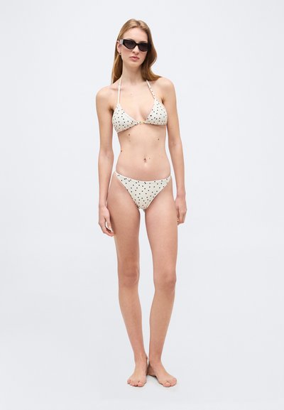 Γυναίκα με λευκό μπικίνι με μαύρες πουά και μαύρα γυαλιά ηλίου, όρθια, ξυπόλητη, μπροστά σε απλό φωτεινό φόντο.