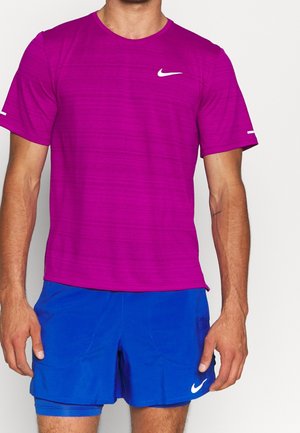 Mężczyzna ubrany w magentową koszulkę sportową Nike z krótkim rękawem oraz niebieskie spodenki sportowe Nike z wbudowanym podkładem kompresyjnym.