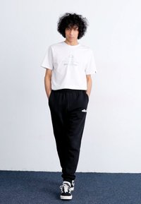 Weißes T-Shirt mit kurzen Ärmeln und bedrucktem Design, kombiniert mit schwarzen Sweatpants mit Logo und schwarzen Turnschuhen mit weißen Sohlen.