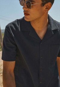 Chemise à manches courtes marine en tissu texturé, avec un bouton de fermeture et une poche sur la poitrine gauche, présentant un col classique.