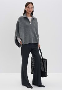 Frau trägt einen grauen Reißverschluss-Poncho, schwarze weit geschnittene Hose, schwarze spitz zulaufende Schuhe und hält eine große schwarze Netz-Totetasche.