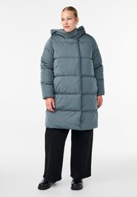 Pūdera jaka attēlā ir nomierinošā teal krāsā, ar kapuci, izšūtu dizainu un garām piedurknēm, apvienota ar brīvām melnām biksēm un izturīgām melnām zābakām.