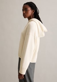 Felpa con cappuccio color crema, lavorata a maglia, con polsini e orlo a costine. Presenta un cappuccio spazioso e una vestibilità comoda, indossata con pantaloni grigi a righe sottili.