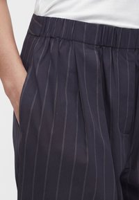 Άτομο φορά σκούρο μπλε παντελόνι με ρίγες και ελαστική ζώνη μέσης, με το χέρι στην πλαϊνή τσέπη, συνδυασμένο με λευκό ριμπ τοπ.