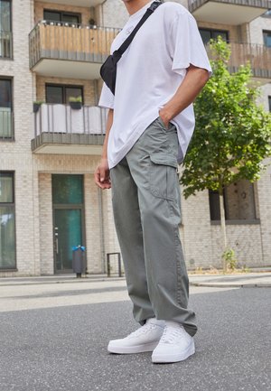 Mann steht im Freien und trägt weiße Sneakers, graue Cargohosen, ein übergroßes weißes T-Shirt und eine schwarze Umhängetasche.