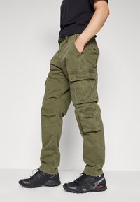 Olivgrüne Cargohosen mit lockerem Schnitt, ausgestattet mit Seitentaschen und verstärkten Nähten. Kombiniert mit schwarzen Sportschuhen.