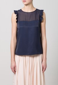 Navy blue silk sleeveless top featuring a sheer upper panel and ruffled shoulder accents, paired with a light pink pleated skirt.