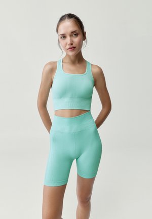 Mujer joven con el cabello recogido, vistiendo un sujetador deportivo y pantalones cortos de ciclista de cintura alta en turquesa claro a juego, de pie con las manos detrás de la espalda.