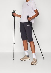 Person som bär en ljuslila t-shirt, svarta shorts, beige sneakers, håller i svarta vandringsstavar med grå handtag och vita strumpor.