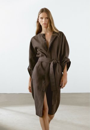 Model walks confidently, wearing a dark brown belted shirt dress with a notched collar, against a minimalistic light background.
