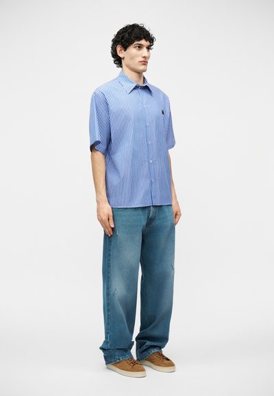 Jeune homme debout, portant une chemise à manches courtes à rayures verticales bleues, un jean bleu ample et des baskets marron sur un fond blanc uni.