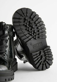 Bottes cheville en cuir verni noir avec lacets décoratifs, semelle épaisse à crampons et détail zippé sur le côté. Présentent une texture brillante.