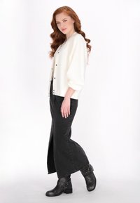 Witte oversized cardigan met zwarte knopen, gecombineerd met een zwart denim rok met hoge taille en een zijsplit, en zwarte enkellaarsjes.