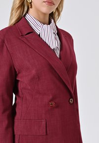 Bordeaux-farbenes, doppelt geknöpftes Blazer mit strukturierten Revers, ausgestattet mit zwei goldenen Knöpfen und einer Seitentasche, über einer gestreiften Bluse getragen.
