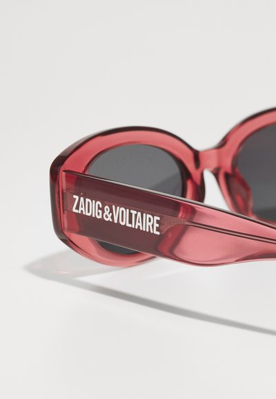 Lunettes de soleil transparentes rouges avec une forme ronde et des verres foncés. Présente le texte blanc "ZADIG&VOLTAIRE" sur les branches. Fini brillant.