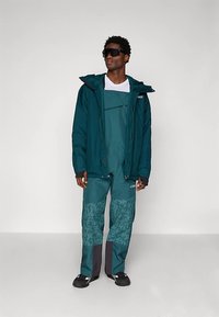 Türkiz színű vízálló dzseki kapucnival, hozzáillő texturált mintás kertésznadrággal rétegezve. Fekete részletek a mandzsettánál és a bokánál.