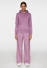 Bluza z kapturem w kolorze lawendowym, z przednią kieszenią i sznurkami, zestawiona z luźnymi spodniami w tym samym kolorze. Oba wykonane z miękkiej, fakturowanej tkaniny.