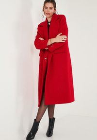Red wool coat with a long silhouette, wide lapels, two front pockets, and a single button closure. Worn with black ankle boots.
