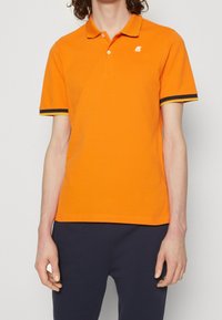 Homme portant un polo à manches courtes orange vif avec des bordures noires et jaunes sur les manches, associé à un pantalon bleu marine foncé, se tenant devant un fond blanc.