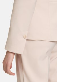 Světlý béžový blejzr s přiléhavým střihem, texturovaná tkanina a jediný krémový knoflík na manžetě. Kalhoty jsou v souladu a mají elegantní střih.