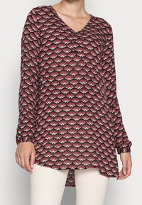 Rode blouse met patroon, voorzien van een V-hals, lange mouwen en een uitlopende onderkant. Heeft een waaier-achtige ontwerp in contrasterende tinten crème en zwart.