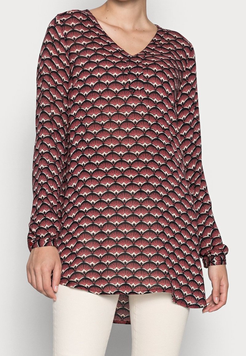 Rode blouse met patroon, voorzien van een V-hals, lange mouwen en een uitlopende onderkant. Heeft een waaier-achtige ontwerp in contrasterende tinten crème en zwart.