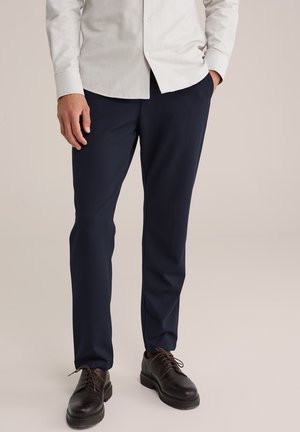 Pantalones marinos con un corte entallado, fabricados en una tela suave. Combinados con zapatos negros de cordones y complementados con una camisa de botones en gris claro.