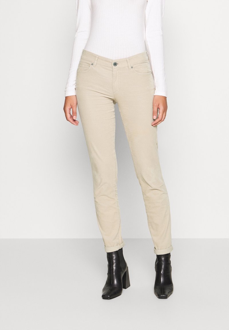 Pantalon beige fuselé en tissu doux, avec poches avant, fermeture par bouton, et ourlet retroussé. Porté avec des bottines noires à talons hauts.