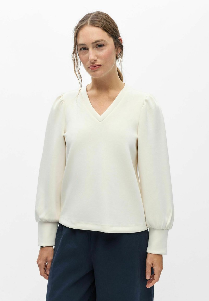 Femme aux cheveux bruns attachés en queue de cheval basse, portant un chemisier blanc à manches longues, col en V, manches bouffantes, et un pantalon bleu foncé sur un fond uni.