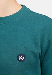 Felpa in cotone color teal con un patch rotondo blu navy ricamato in bianco, colletto a costine e texture liscia.