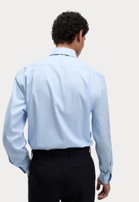 Jasnoniebieska koszula z długim rękawem, prosta pod kolanem, gładka w dotyku, z mankietami na guziki, zestawiona z ciemnymi spodniami.