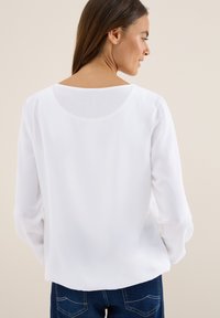 Blouse blanche à manches longues avec un décolleté large, fabriquée en tissu léger, présentant une texture lisse et une coupe décontractée, vue de dos.