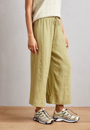 Person trägt hellgrüne, weit geschnittene Hose mit Kordelzug in der Taille und beige Sneakers mit schwarzen Akzenten, steht auf einem Holzboden.