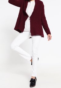 Bordeaux gebreide cardigan over een witte blouse en slim fit witte broek. Zwarte schoenen met een strikaccent maken de outfit compleet.