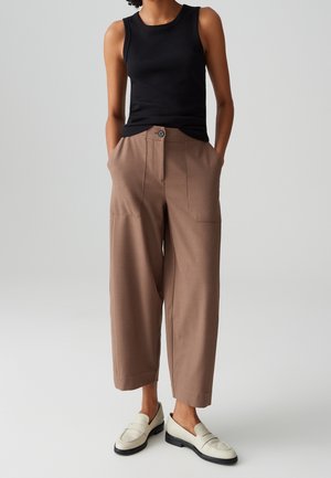 Person wearing a black sleeveless top, loose tan cropped pants with pockets, and white slip-on loafers standing with hands in pockets.