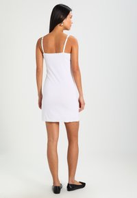 Abito da notte bianco con pizzo, spalline regolabili, lunghezza sopra il ginocchio, silhouette aderente, texture liscia, abbinato a scarpe nere basse.