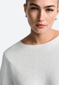 Light gray textured sweater with a wide neckline. The fabric has a subtle sheen, complementing the model's clear blue eyes and simple silver earrings.