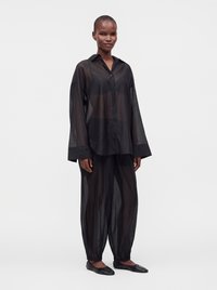 Personne portant une chemise noire ample, transparente et boutonnée ainsi qu'un pantalon transparent assorti, debout avec une posture détendue et des ballerines noires sur un fond uni.