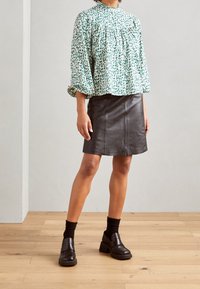 Bluse mit Blumenmuster in Grün und Weiß, mit Puffärmeln und hohem Ausschnitt, kombiniert mit einem schwarzen Leder-Minirock und klobigen Absatzschuhen.