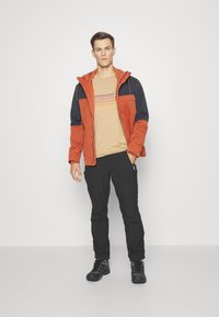 Giacca da esterno in tessuto arancione e blu scuro, con cappuccio, cerniere e tasche, abbinata a una camicia a righe beige, pantaloni neri e stivali.