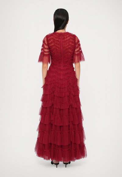 Raudona sluoksniuota suknelė, pagaminta iš tekstūruoto audinio, turinti trumpas permatomas rankoves ir prigludusį liemenį. Sijonas turi kelis volanų sluoksnius.