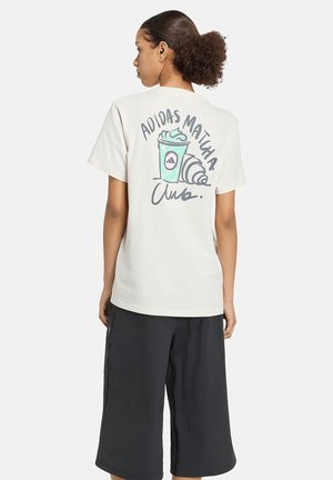 Kobieta z kręconymi włosami w białym t-shircie z grafiką napoju matcha i rogalika oraz czarnych szerokich spodniach, stoi tyłem.