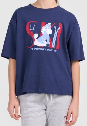 T-shirt bleu marine avec un graphique de caniche skieur, skis, bâtons et texte "SKI It's powder day", porté avec un pantalon de survêtement gris clair.