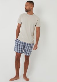 Camiseta de algodón color beige claro combinada con bañador de cuadros azul y púrpura. El bañador tiene una cinturilla elástica y bolsillos laterales.