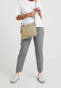 Beżowa torba na ramię z teksturowanego materiału, zamek błyskawiczny i regulowany pasek. Noszona z szarymi spodniami w kratkę i białymi trampkami.