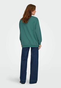 Felpa verde scuro con polsini e orlo a costine, abbinata a jeans scuri a vita alta. Il tessuto appare morbido con una vestibilità comoda.