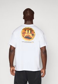 Hvit bomull t-skjorte med en sirkulær grafikk som har et basketballdesign i oransje og gult, tekst i svart og gull, på baksiden.