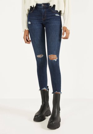 Jean slim fit bleu foncé déchiré associé à des bottines noires épaisses à plateforme et à un pull court blanc effet usé.
